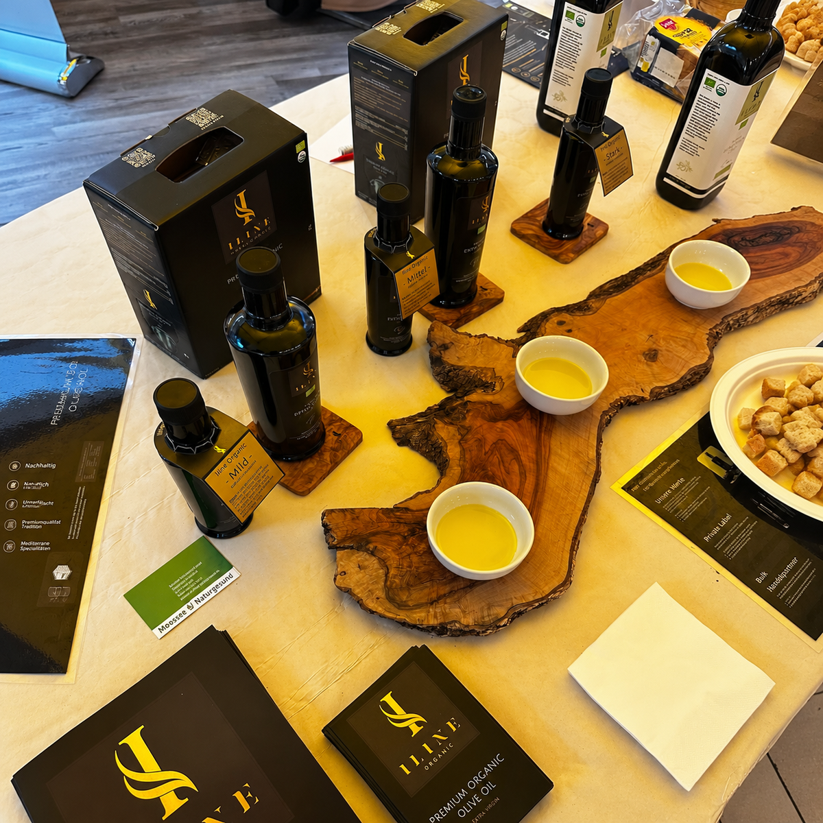 Iline Organic Degustationstisch mit Bio-Olivenölflaschen, Produktboxen und Probeschalen auf Olivenholzbrettern sowie Brot für die Verkostung.