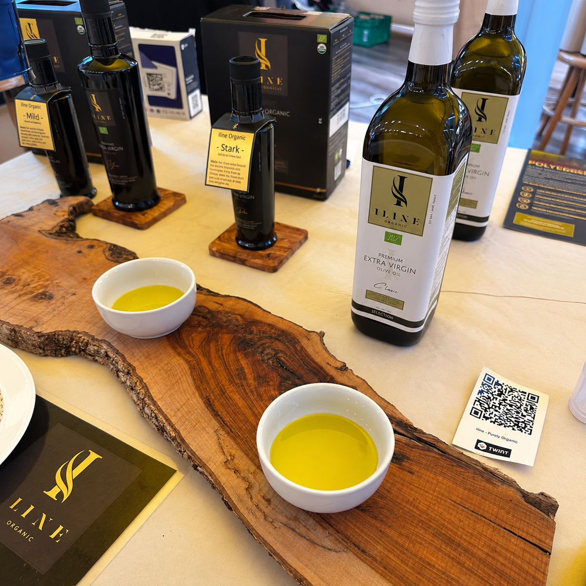 Iline Organic Classic Bio-Olivenöl in einer grossen Flasche neben zwei mit Olivenöl gefüllten Probeschalen auf einem Olivenholzbrett.