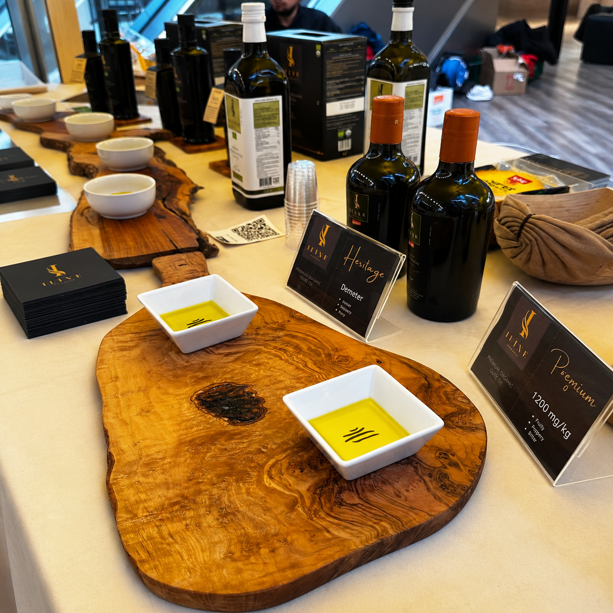 Iline Organic Olivenöl-Degustation mit Heritage- und Polyphenol-Produktkarten, dunklen Olivenölflaschen und zwei Probeschalen auf Olivenholz.