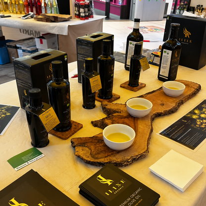 Verschiedene Iline Organic Bio-Olivenöle und Produktverpackungen neben drei Probeschalen auf rustikalen Olivenholzbrettern.